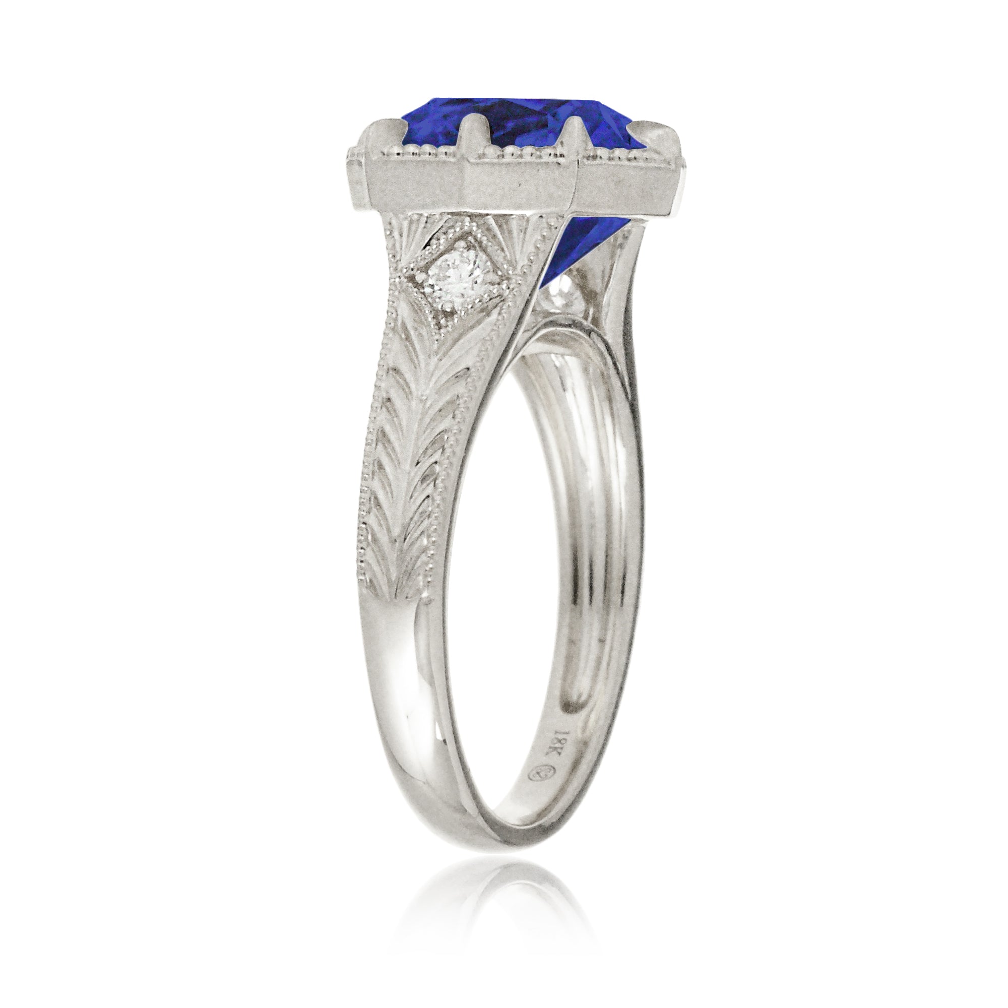 A cushion cut blue sapphire glimmers at the heart of this vintage-inspired engagement ring, set in polished white gold. The detailed band features hand-carved artistry, enhancing the antique character and timeless appeal.